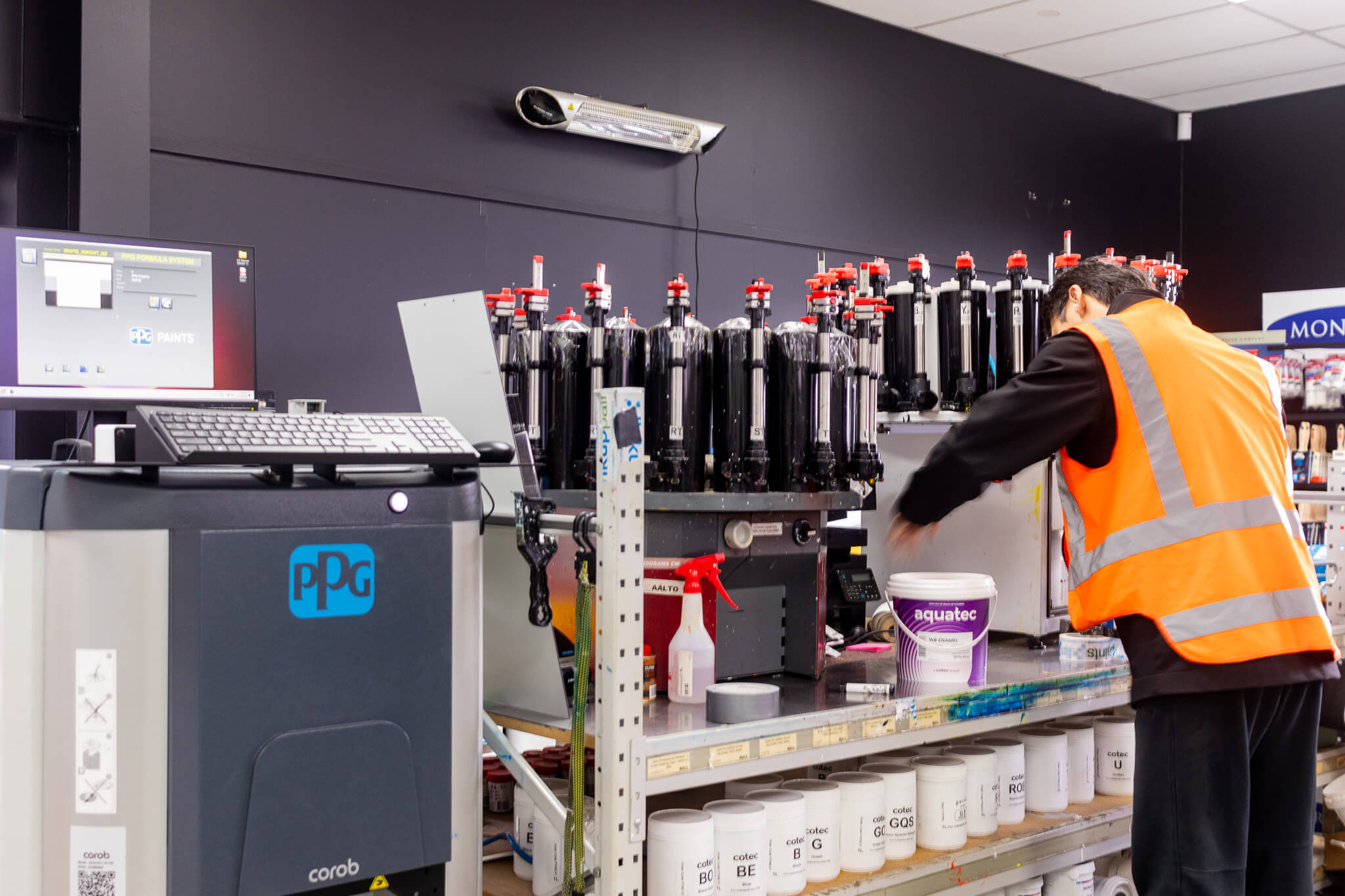 A person placing a label on a pail of mixed paint, surrounded by tinting carousels and automatic tint machines